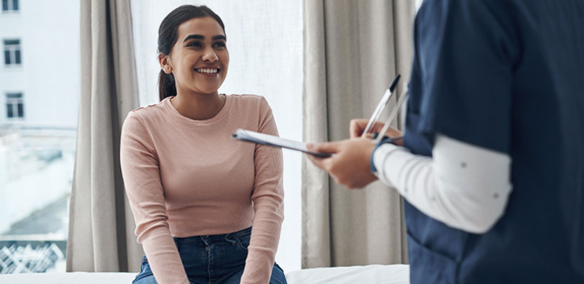 Woman sitting on a medical exam table smiling at a healthcare professional holding a clipboard.