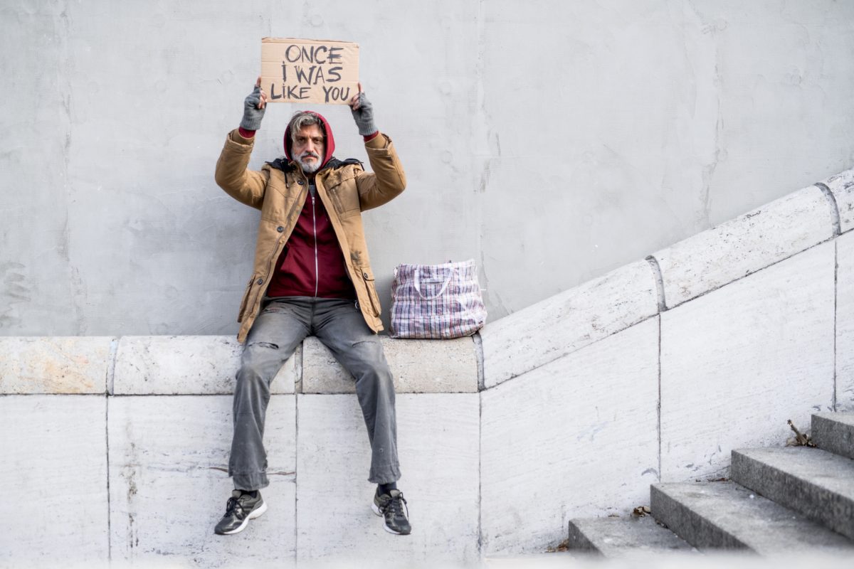 Homeless beggar man sitting outdoors in city holding will work for food cardboard sign above his head, asking for money donation.